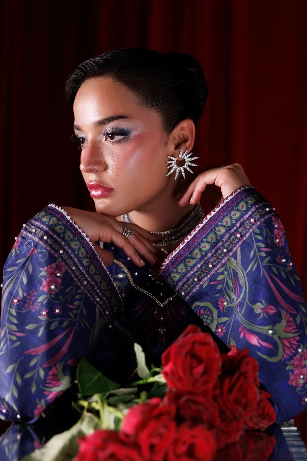 A woman with dark hair styled in an updo wears a dark purple embroidered blouse with pink and green floral patterns, silver jewelry, and a starburst earring, holding a bouquet of red roses against a dark red curtain background.