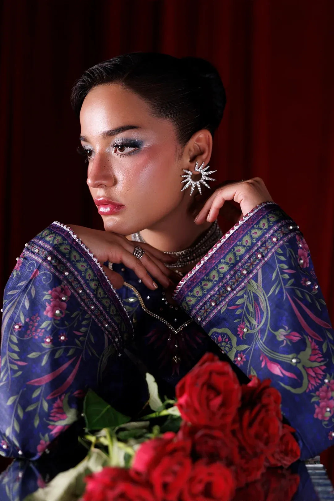 A woman with dark hair styled in an updo wears a dark purple embroidered blouse with pink and green floral patterns, silver jewelry, and a starburst earring, holding a bouquet of red roses against a dark red curtain background.