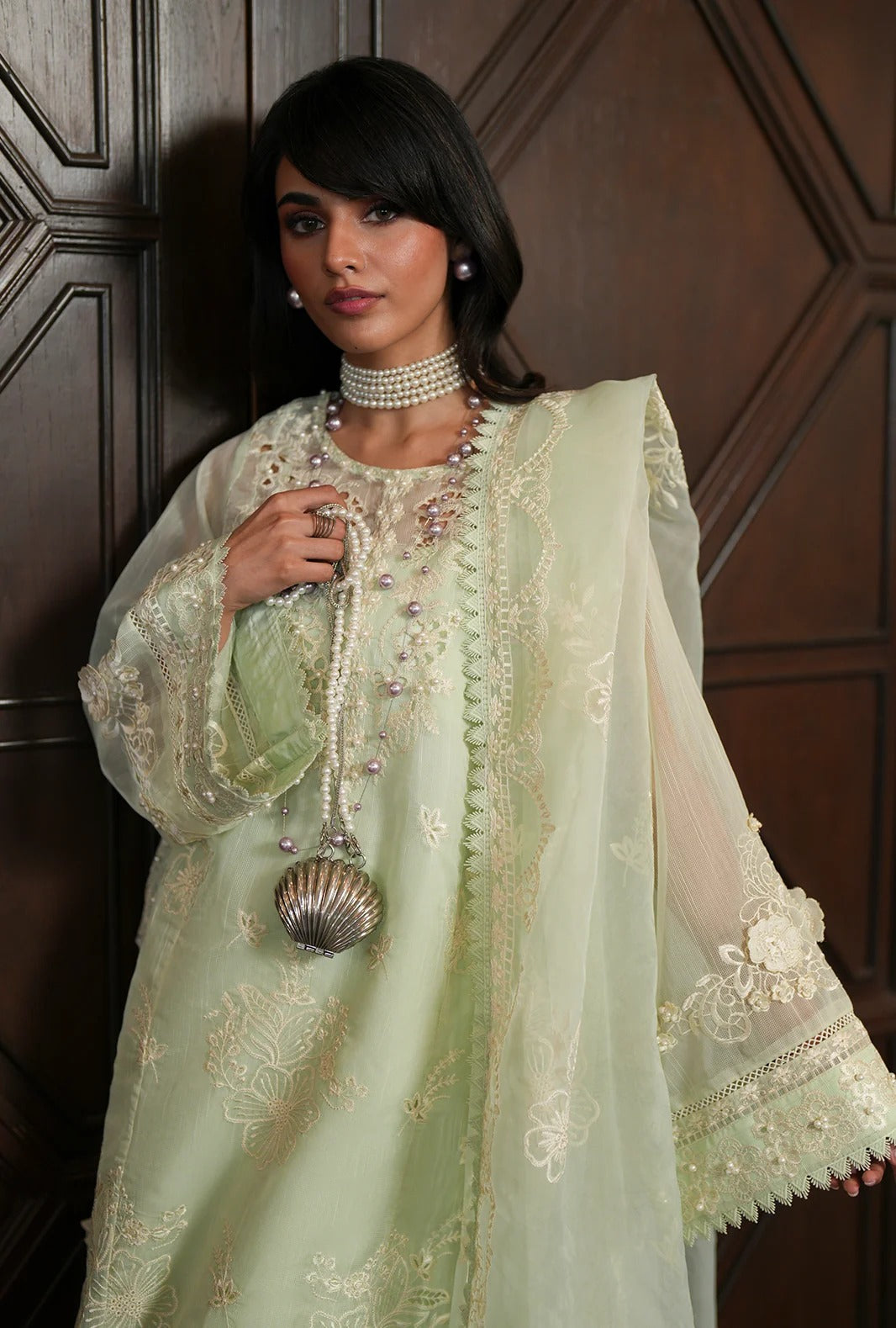 A woman models a traditional light green embroidered outfit with a matching sheer dupatta, pearl choker, and a metallic shell-shaped pendant, posed against a dark wood paneled background.