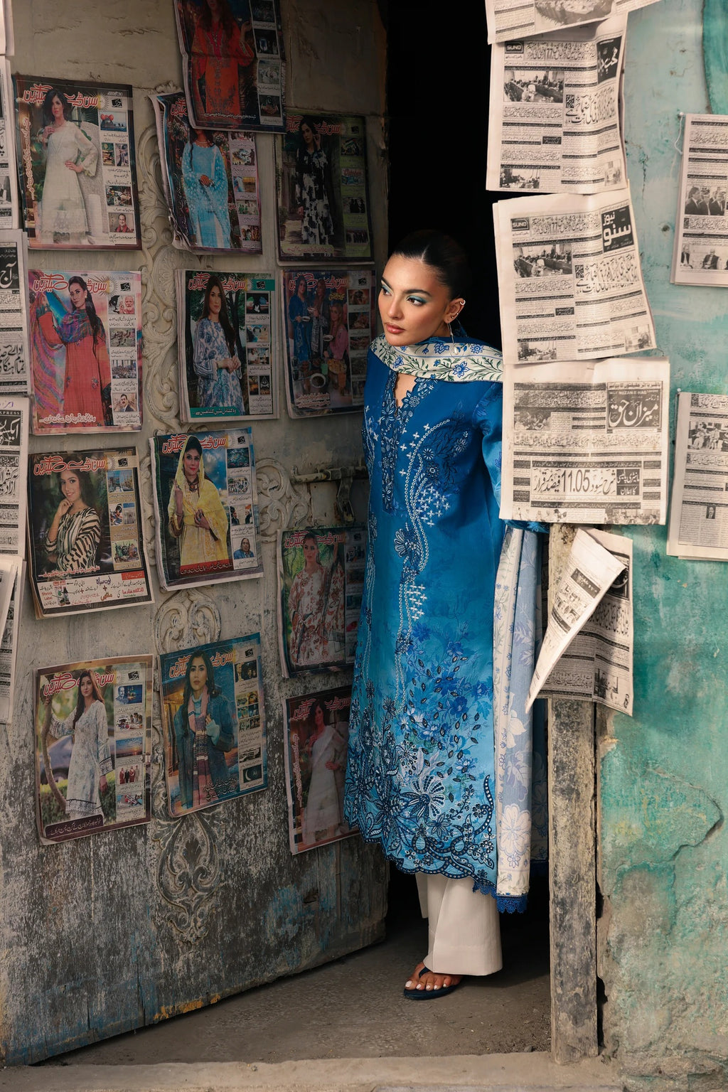 A woman wearing a vibrant blue embroidered traditional outfit with white pants stands in a doorway surrounded by vintage posters and newspaper clippings.