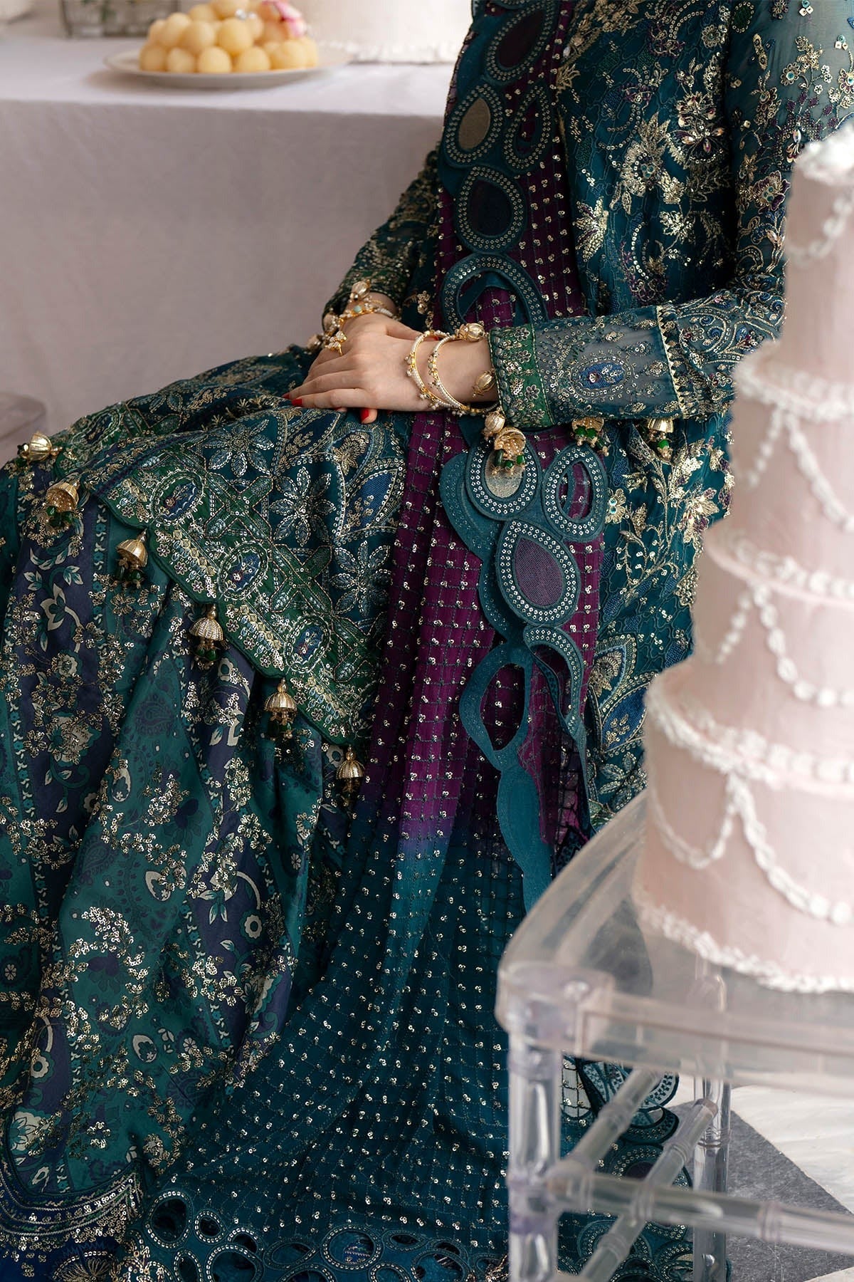 A close-up of a person wearing an ornate traditional outfit in deep teal and purple with intricate gold embroidery, seated indoors near a table and a pink tiered cake on a clear acrylic stand.