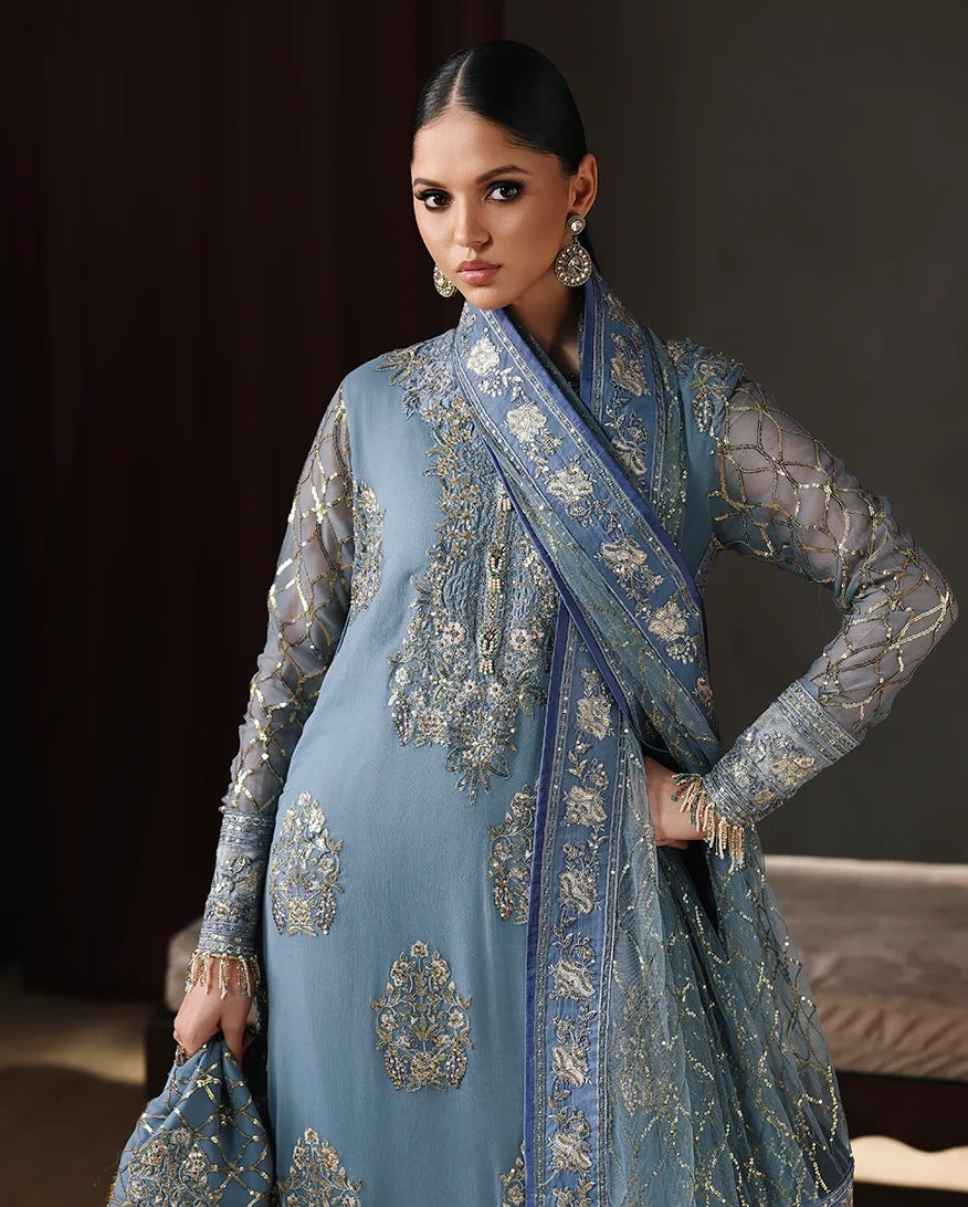 A woman models a light blue traditional embroidered outfit with sheer sleeves and ornate metallic embroidery, paired with a matching draped scarf, in a dark indoor setting.