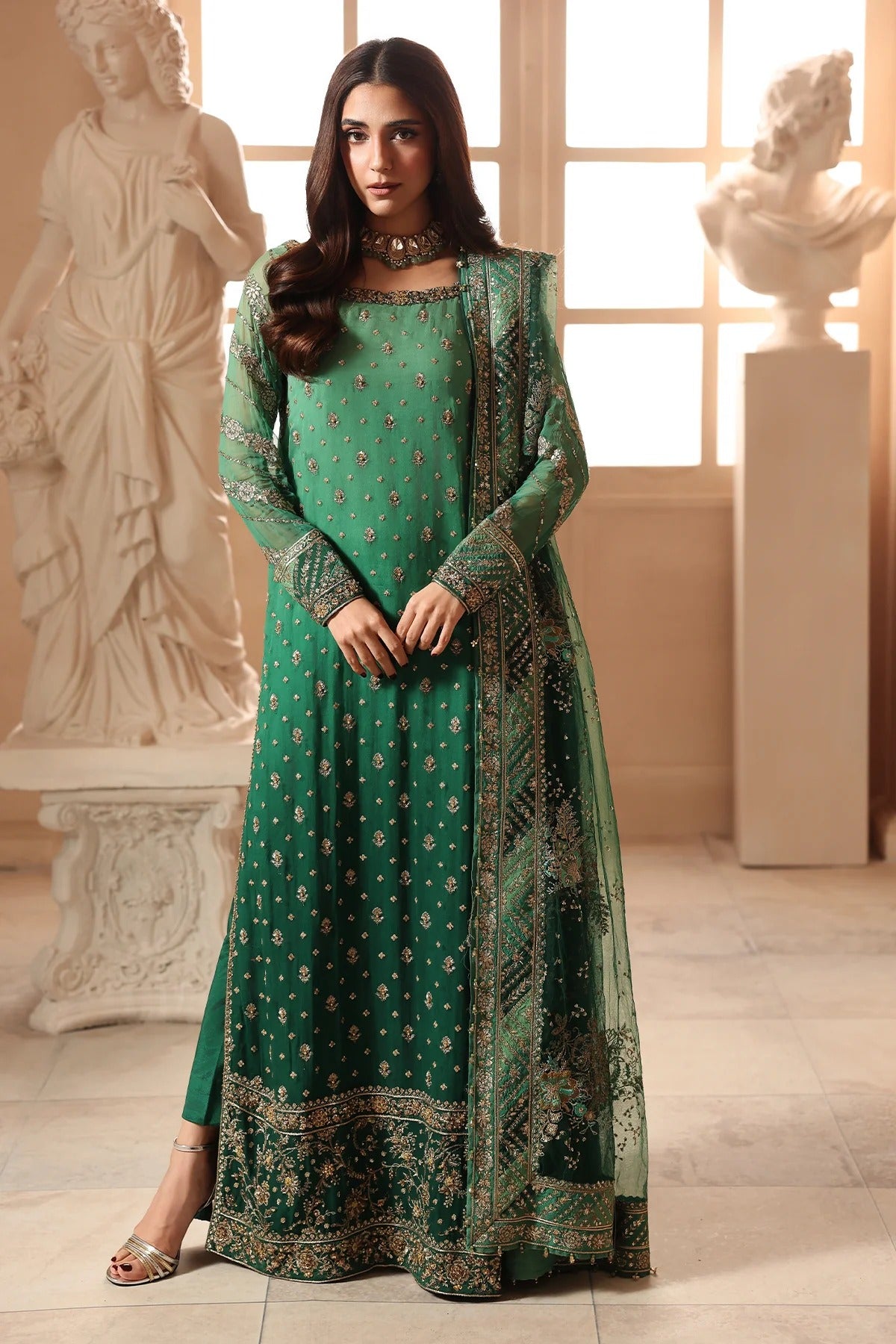 A woman models a traditional green ethnic outfit with gold embroidery and a matching sheer dupatta in an indoor setting with classical bust statues.