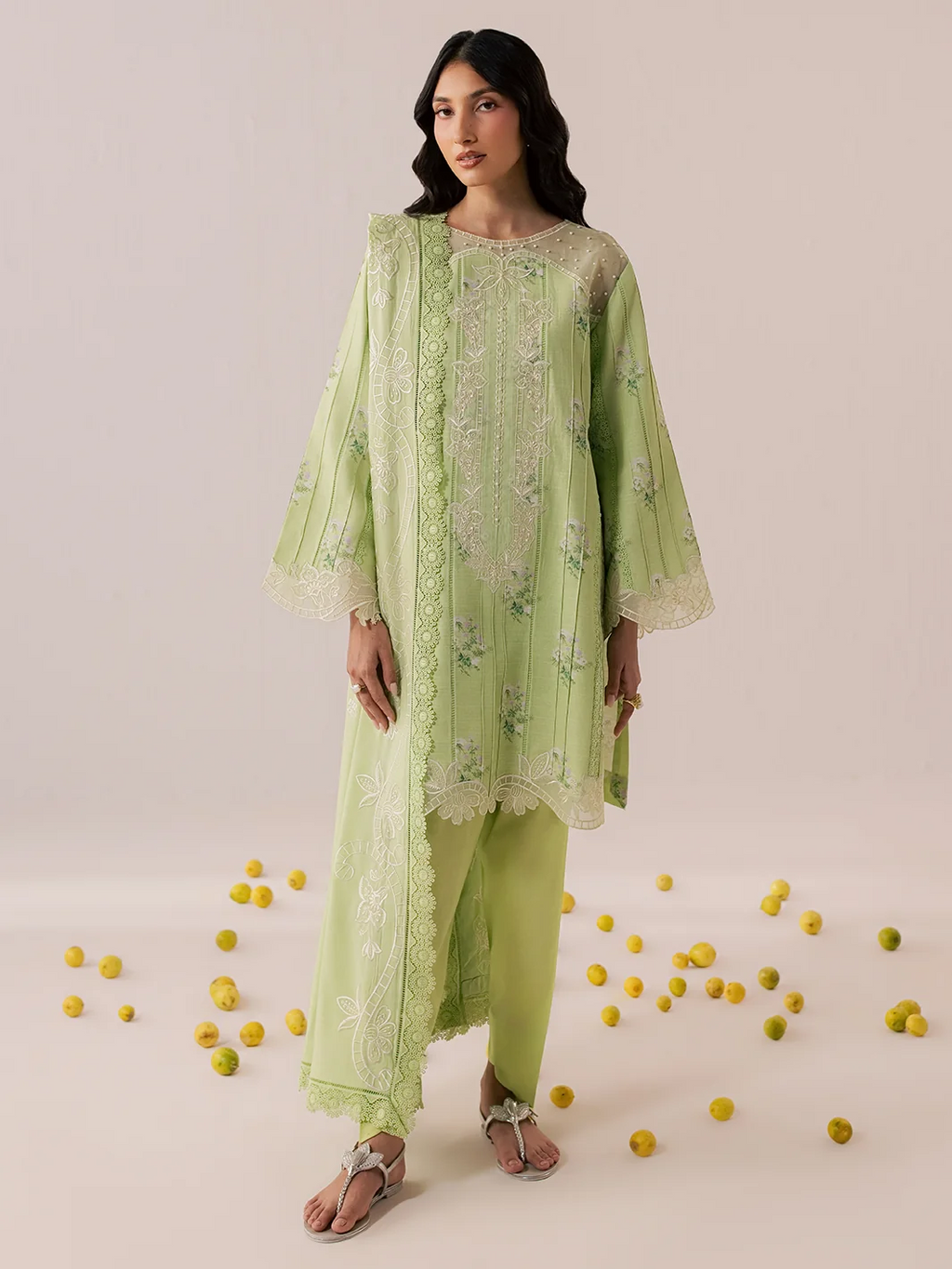 A woman models a light green three-piece traditional outfit with matching embroidered sheer jacket and pants, standing against a plain beige studio backdrop with scattered yellow pom-poms on the floor.