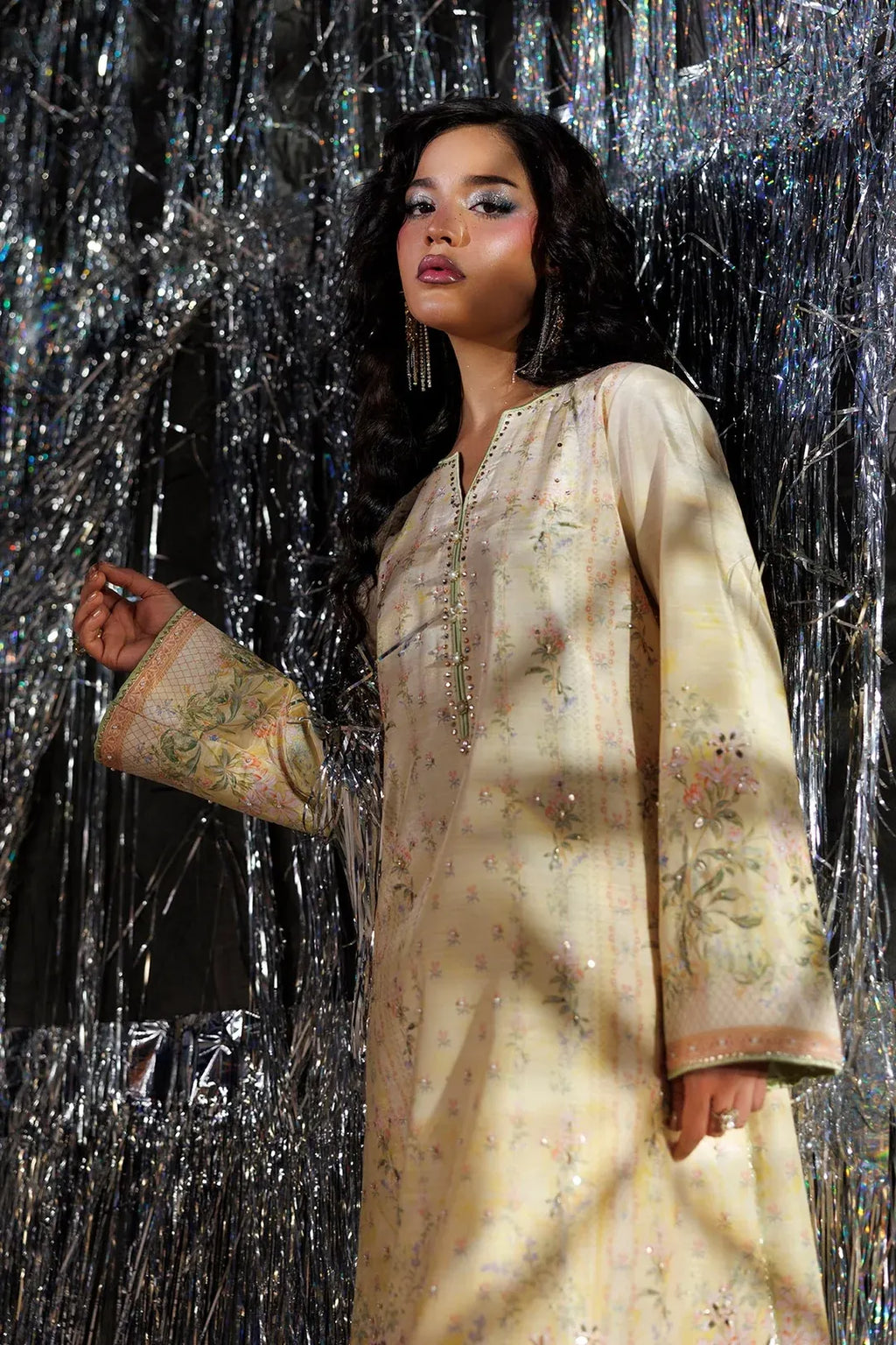 A woman wearing a cream-colored embroidered traditional long-sleeve dress with metallic embellishments, standing in front of a shiny silver tinsel backdrop.
