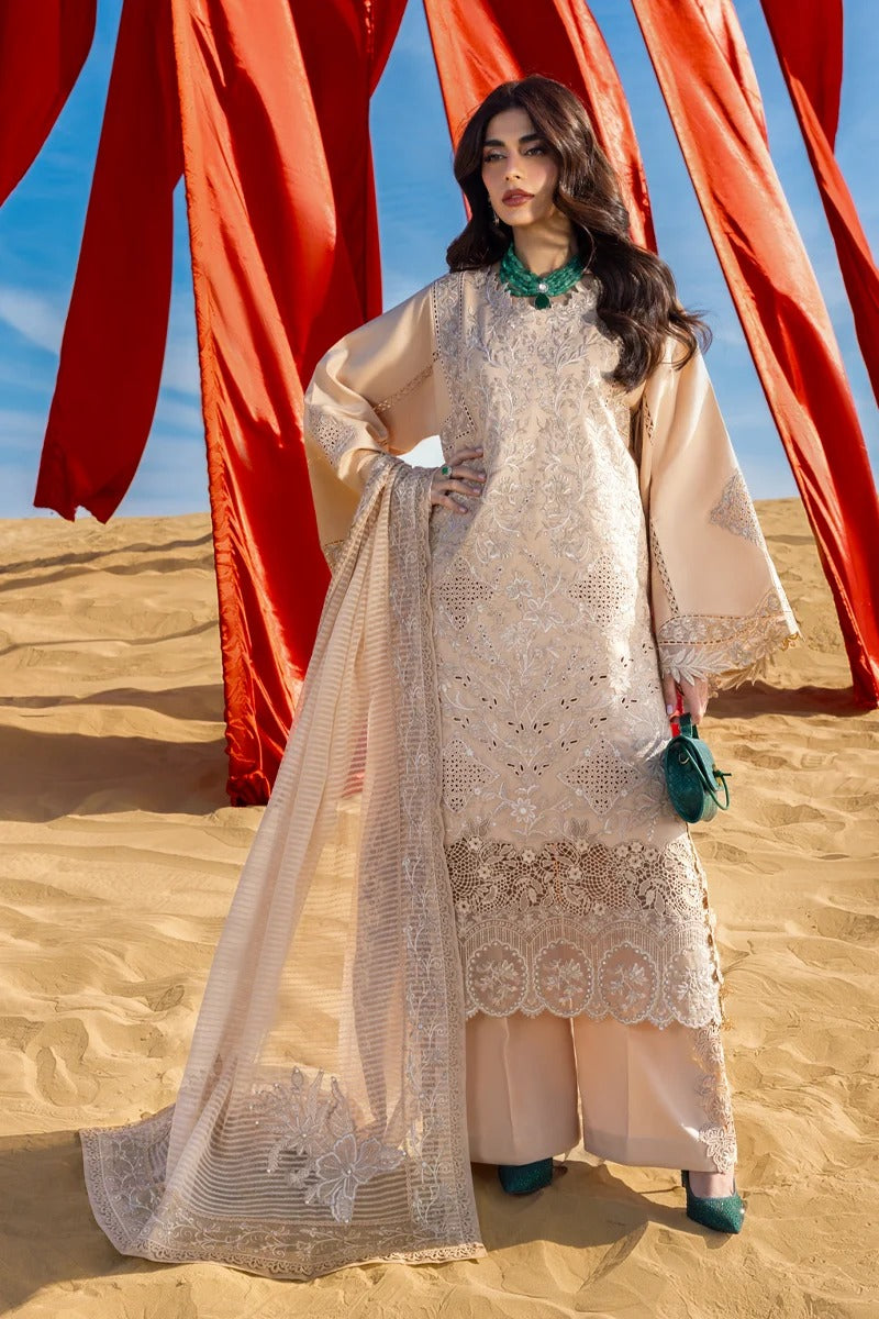 A woman stands in a sandy desert setting wearing a cream-colored embroidered traditional outfit with wide sleeves and a matching sheer dupatta. She is posed in front of large red fabric drapes under a blue sky.