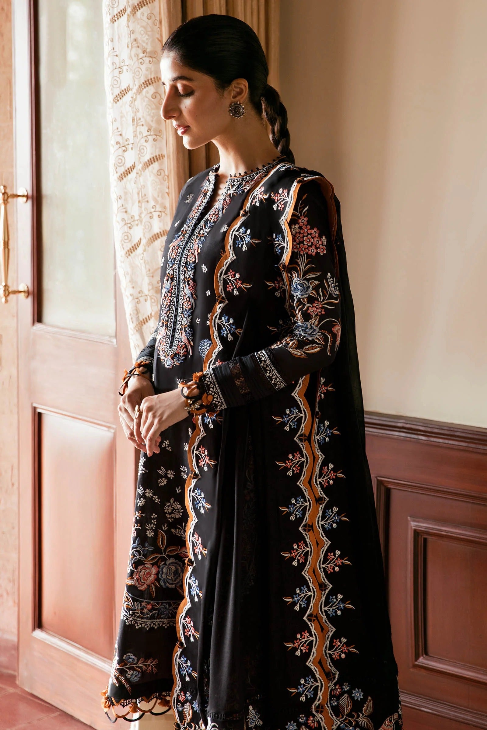 A woman wearing a black embroidered traditional outfit with floral detailing in beige, gold, and blue, standing indoors near a window with curtains and wooden paneling.
