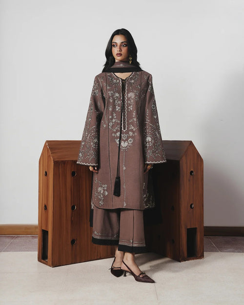 A woman models a brown embroidered kurta and matching wide-leg trousers with black trim, standing in front of a wooden display case against a plain light gray wall.