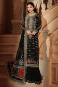 A woman models a black three-piece traditional outfit with extensive silver and gold embroidery, floral prints, and a matching dupatta, posed in front of a grand indoor staircase.