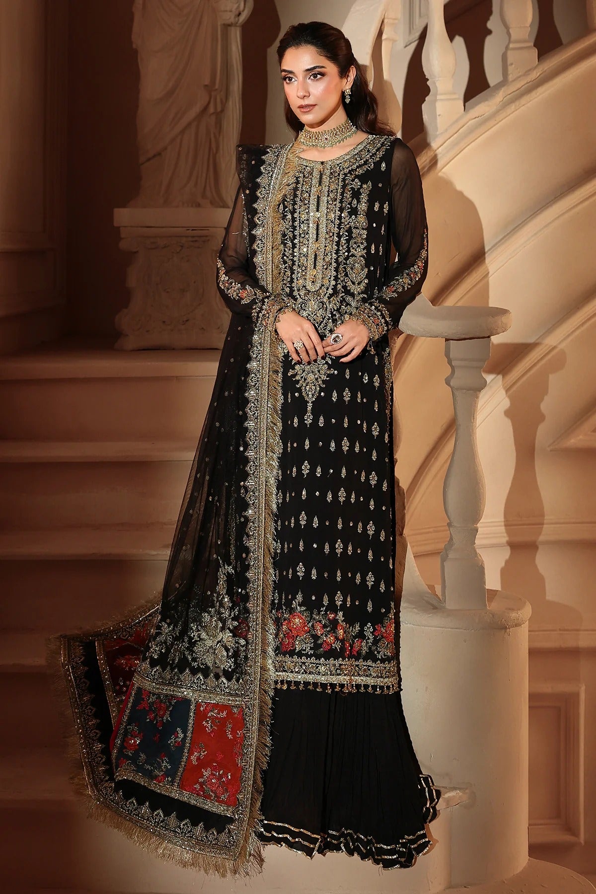 A woman models a black three-piece traditional outfit with extensive silver and gold embroidery, floral prints, and a matching dupatta, posed in front of a grand indoor staircase.