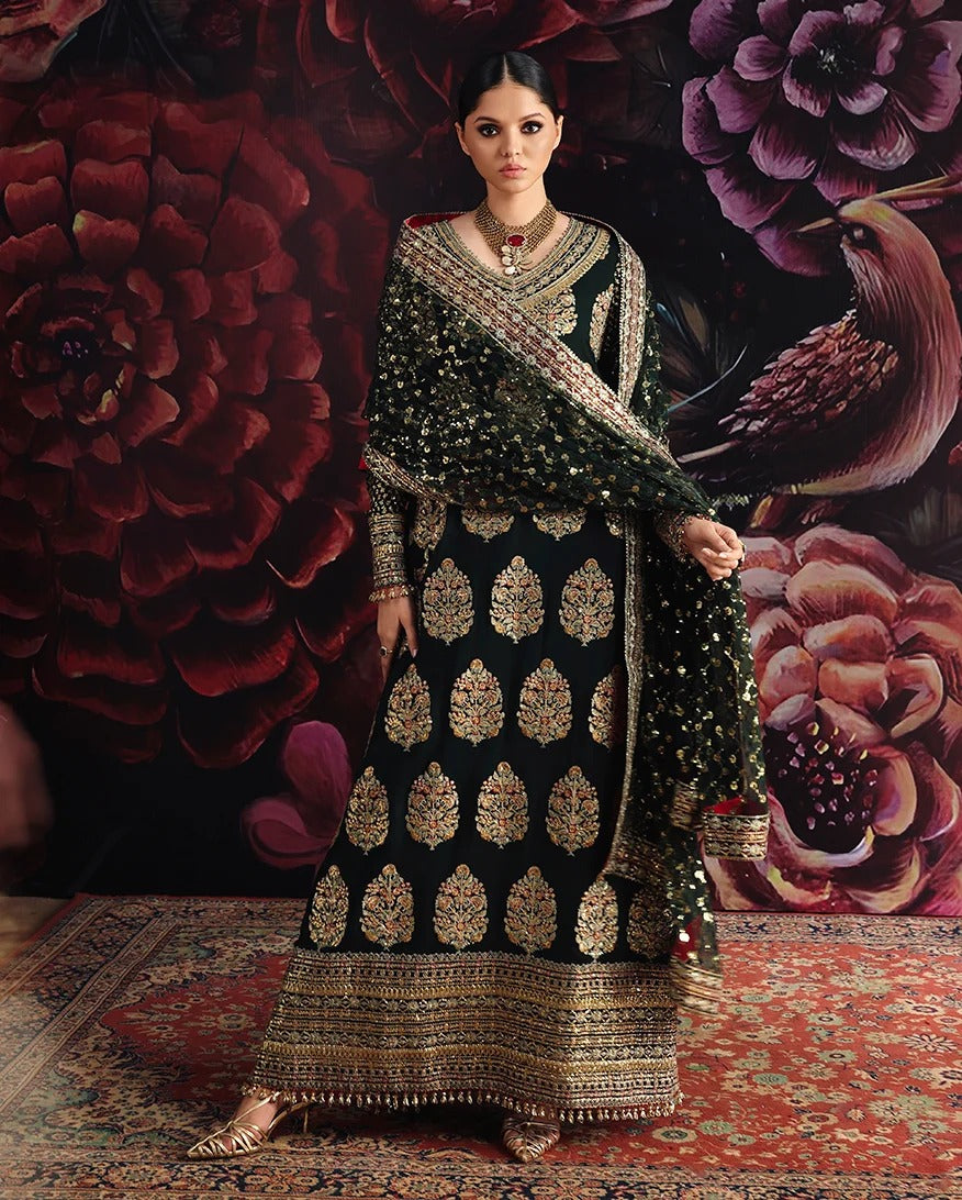 A woman models a traditional black and gold embroidered ethnic outfit with a matching dupatta, set against a dark floral backdrop and patterned rug.