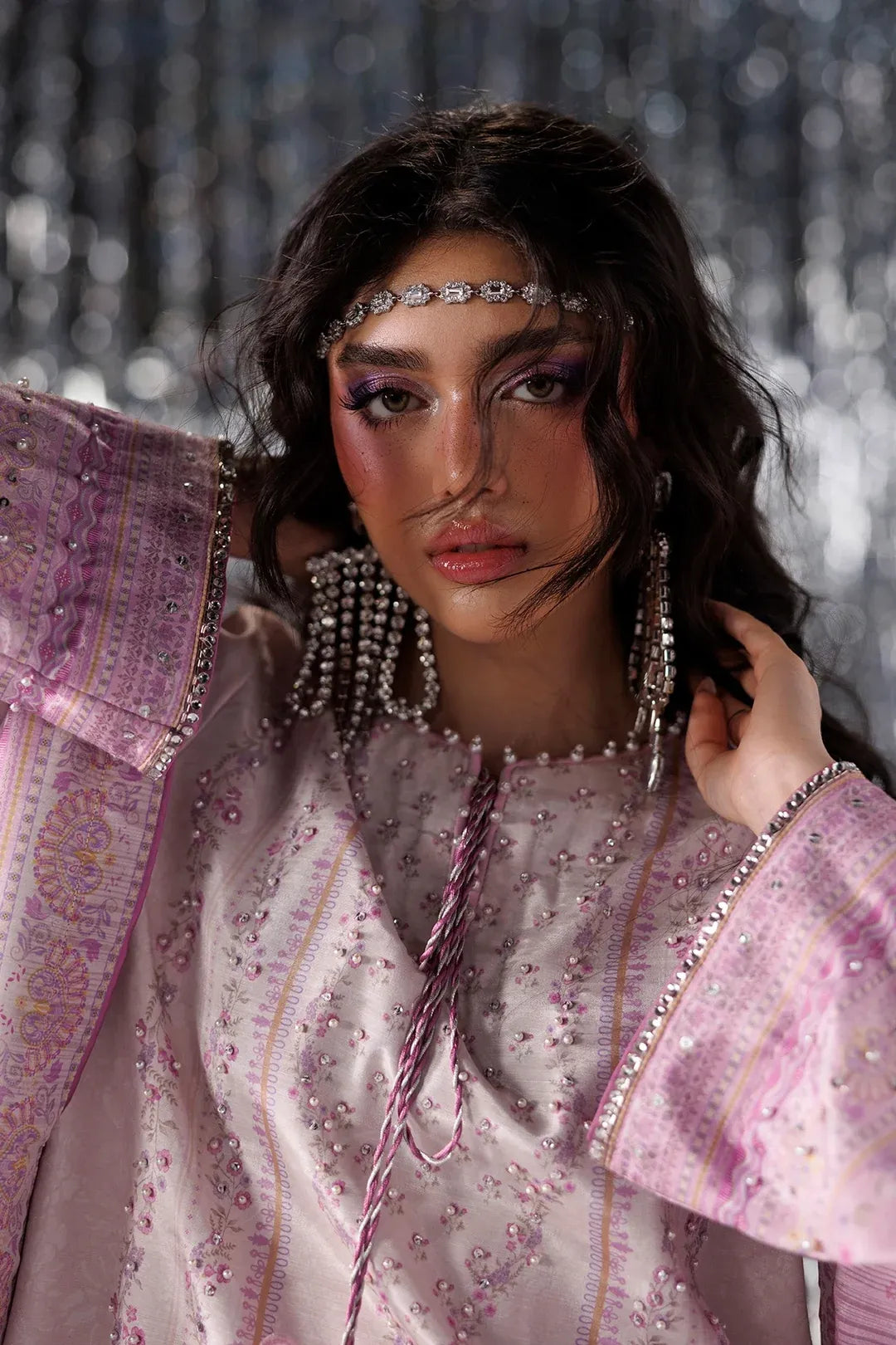 A woman wearing an ornate light pink embroidered traditional outfit with metallic embellishments, matching headpiece, and long dangling earrings, posed against a shimmering silver backdrop.
