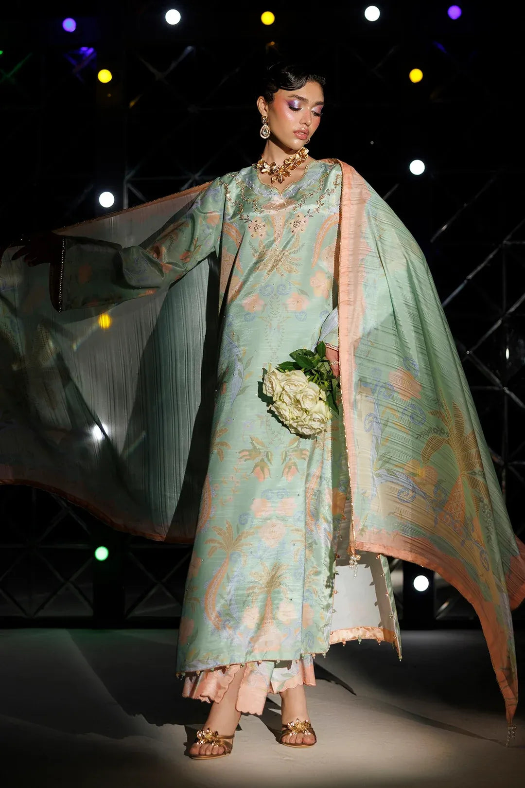 A model walks a fashion runway wearing a mint green printed traditional outfit with a peach dupatta and holding a bouquet of white flowers against a dark stage background with colored spotlights.