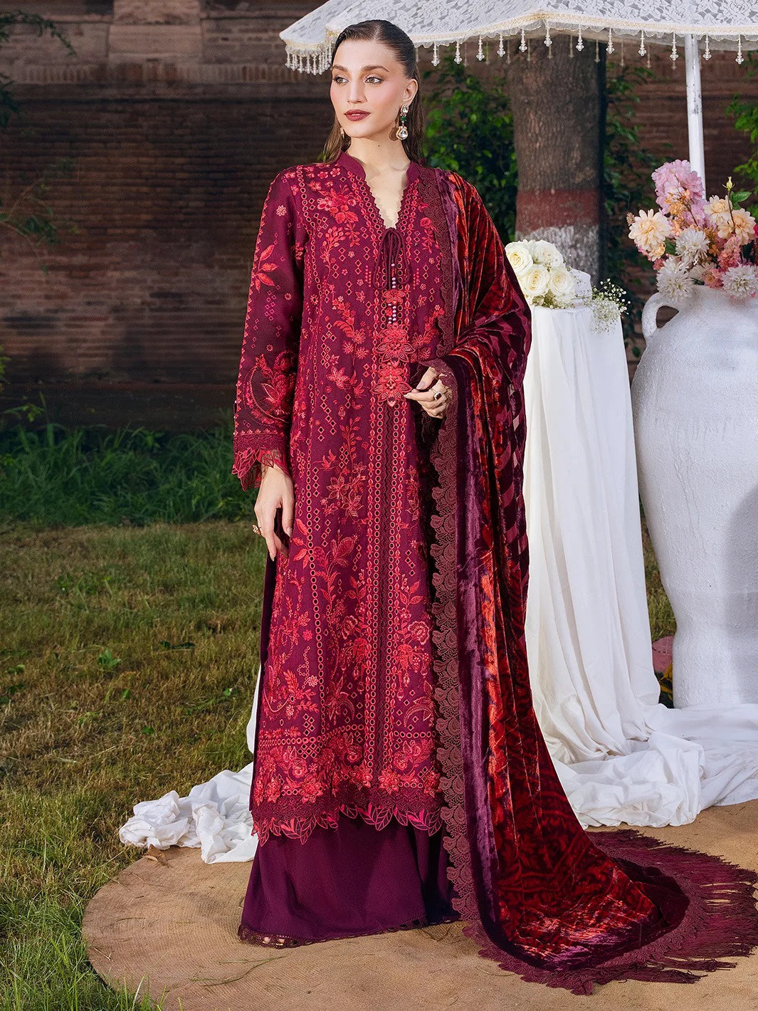 A woman models a traditional three-piece salwar kameez suit in deep maroon and red, featuring intricate floral embroidery, a matching dupatta, and a solid maroon bottom. The setting is an outdoor garden with a white draped backdrop, flowers, and a wooden wall.