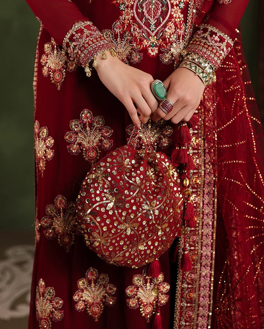 A close-up of a person wearing an ornate deep red embroidered traditional outfit with gold detailing, holding a matching red embellished handbag with tassel handles. The person is also wearing multiple rings and bangles.