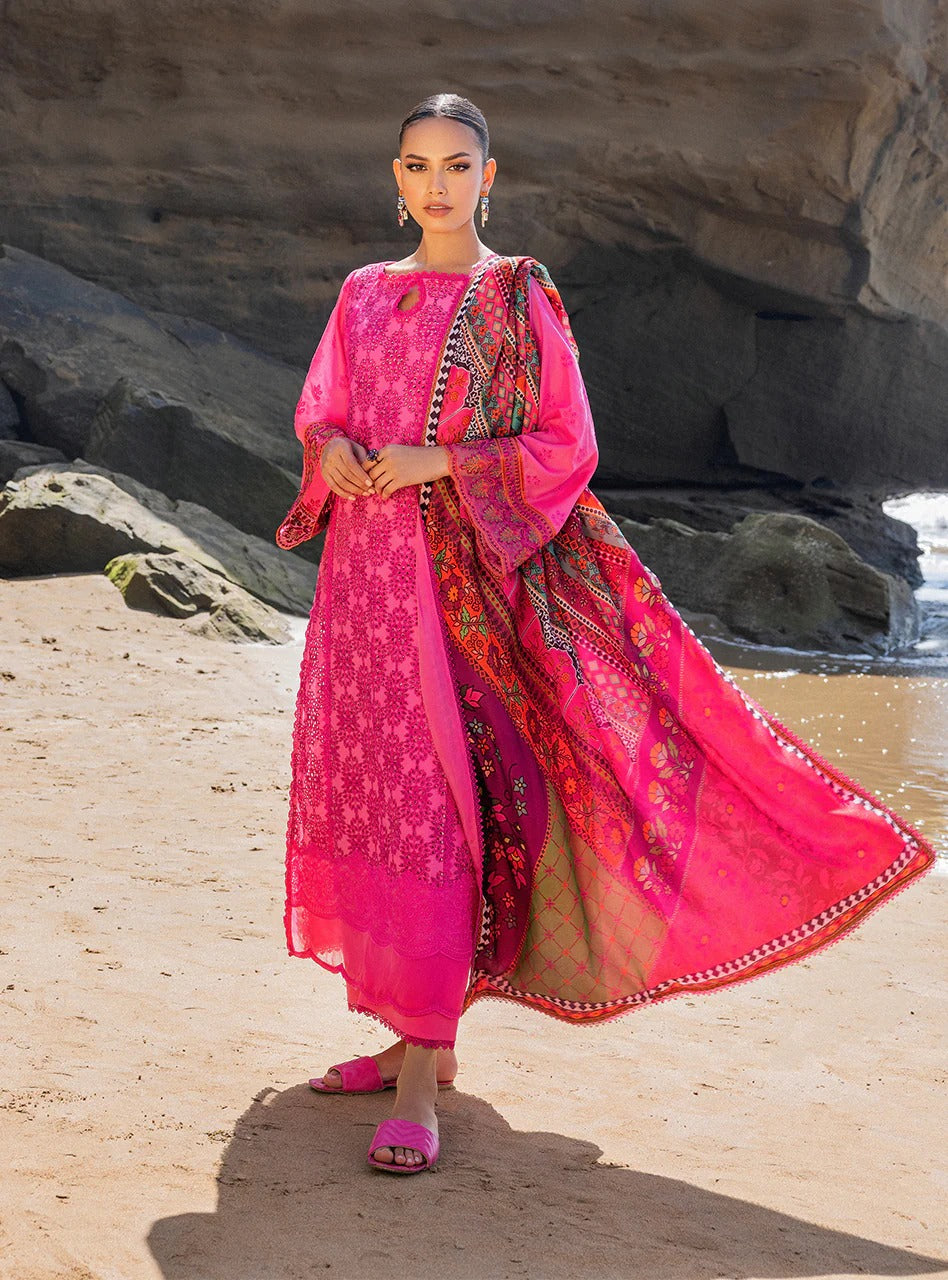 A woman stands on a sandy beach wearing a bright pink traditional outfit with a matching dupatta. The outfit features intricate floral patterns and colorful embroidery, with a rocky shoreline and ocean in the background.