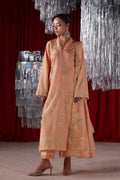 A woman models a peach-colored embroidered traditional outfit with matching pants, standing in front of a red curtain and silver hanging decorations.