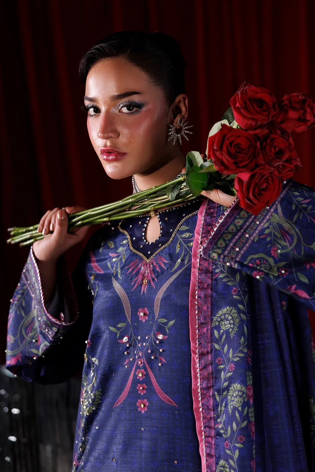 A woman wearing a dark blue embroidered traditional outfit with pink and green floral patterns, holding a bouquet of red roses against a dark red curtain background.