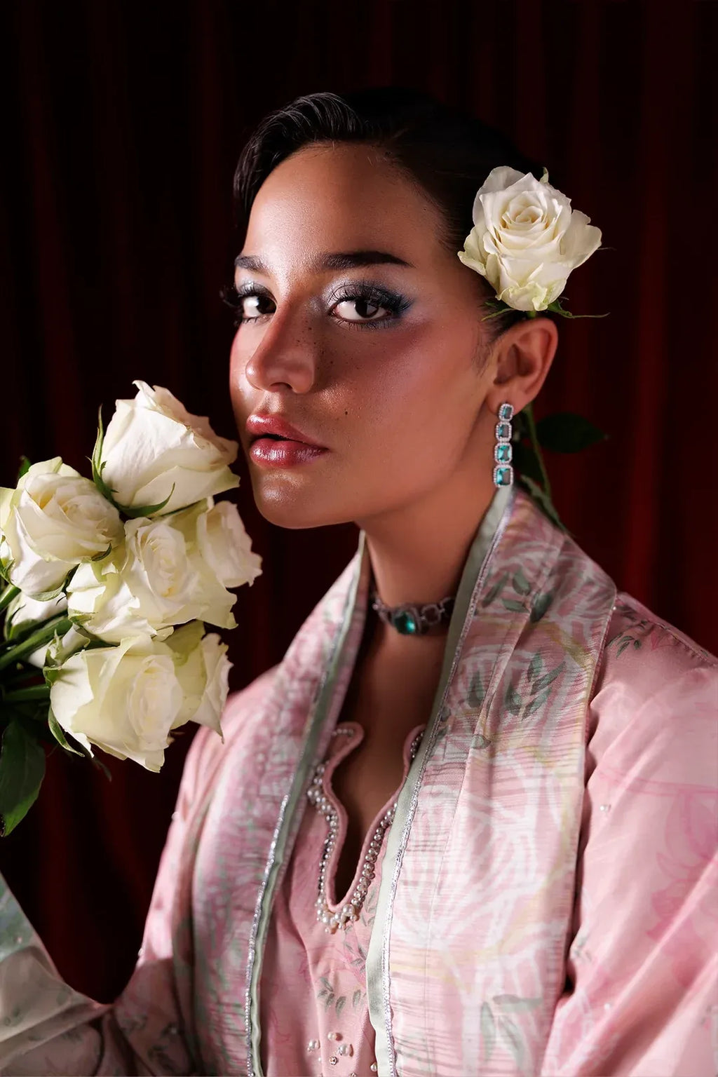 A woman in a light pink patterned traditional outfit with a matching scarf, holding a bouquet of white roses, with a white rose tucked in her hair against a dark red curtain background.