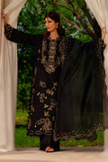 Woman in a black embroidered traditional outfit standing outdoors with greenery in the background.