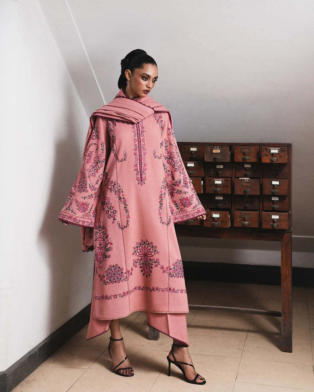 A woman models a pink embroidered traditional outfit with a matching dupatta, featuring intricate floral and paisley patterns, standing in a room with a wooden storage cabinet in the background.