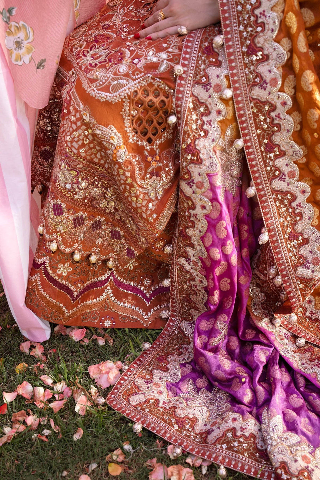 Close-up of an ornate traditional embroidered garment laid outdoors on grass with scattered flower petals. The fabric is richly decorated with metallic embroidery, beadwork, and a contrasting magenta-purple border with white pearl-like accents.