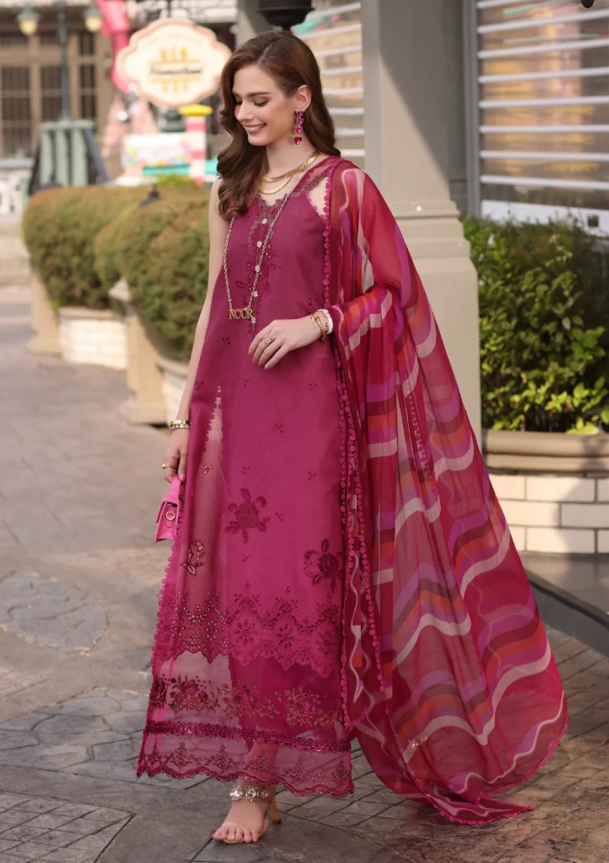 A woman models a magenta pink traditional Indian salwar kameez suit with floral embroidery and a matching striped dupatta in an outdoor urban setting.