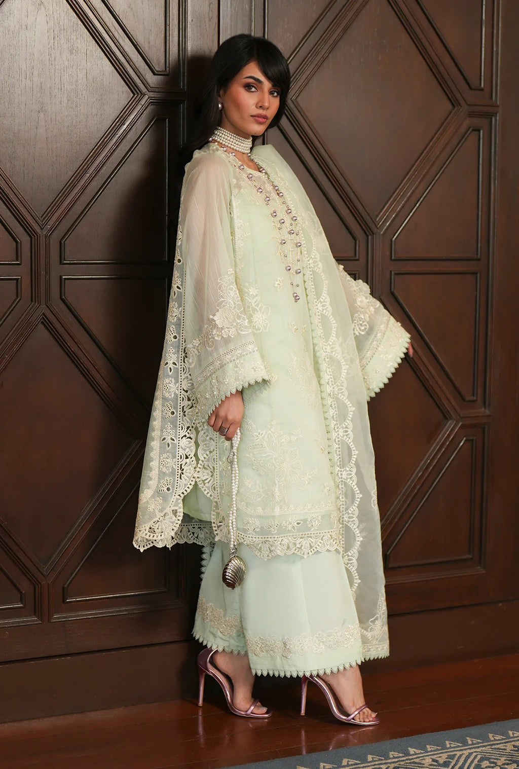 A woman models a traditional light mint green three-piece suit with intricate white embroidery and lace details, standing against a dark brown wooden paneled wall.