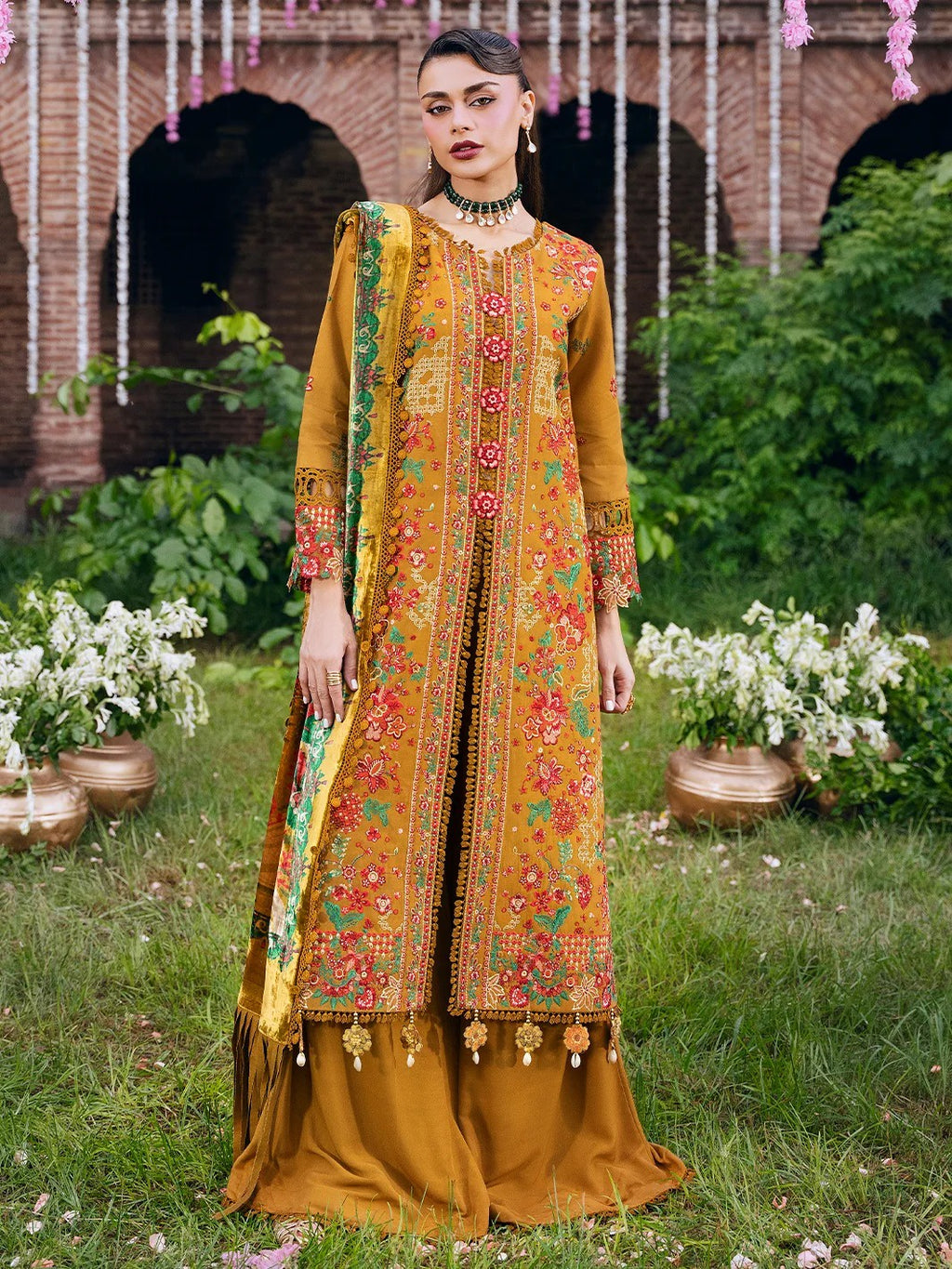 A woman stands outdoors in a garden setting wearing a traditional embroidered outfit. The outfit is a mustard yellow three-piece ensemble with intricate floral embroidery in red, pink, and green, and a matching dupatta draped over one shoulder.