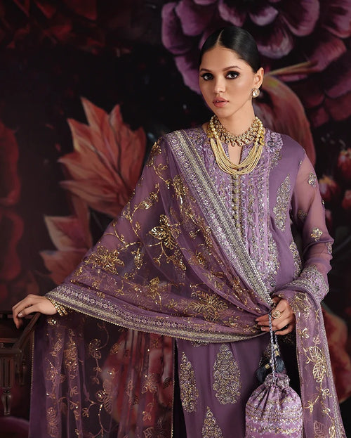 A woman models a purple embroidered traditional outfit with a matching sheer dupatta and gold embellishments, posed against a dark floral backdrop.