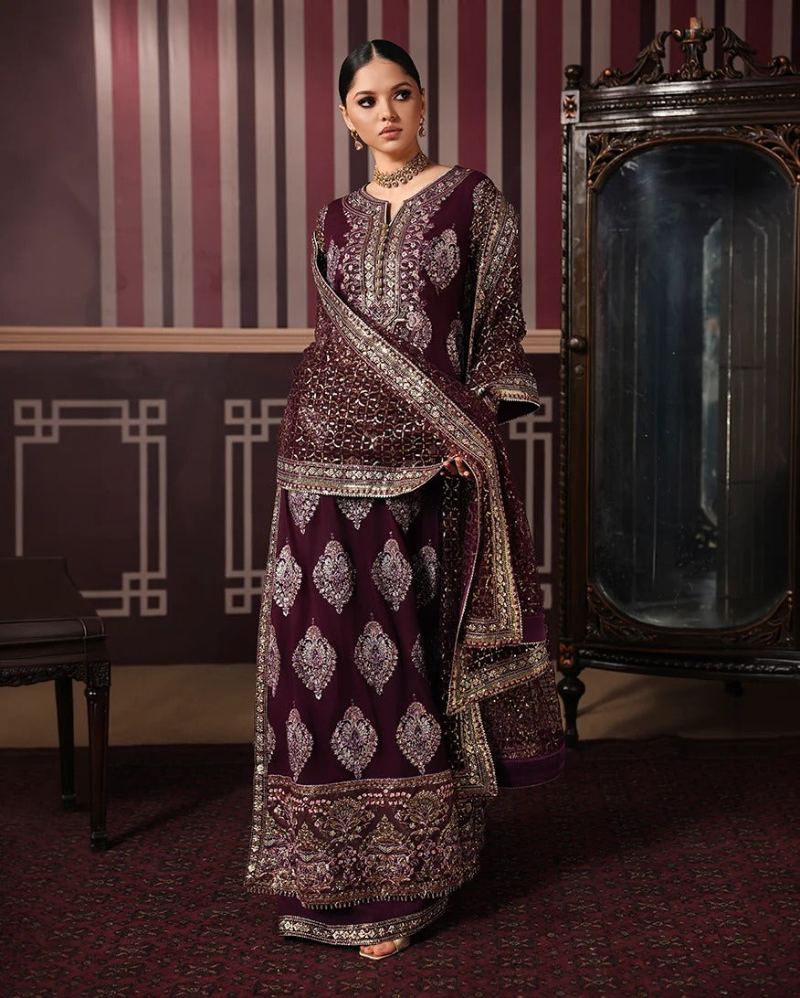 A woman models a traditional Indian outfit consisting of a deep maroon kurta and matching skirt with intricate gold embroidery, paired with a matching embroidered dupatta, set against a vintage-style room background with striped walls and an antique cabinet.