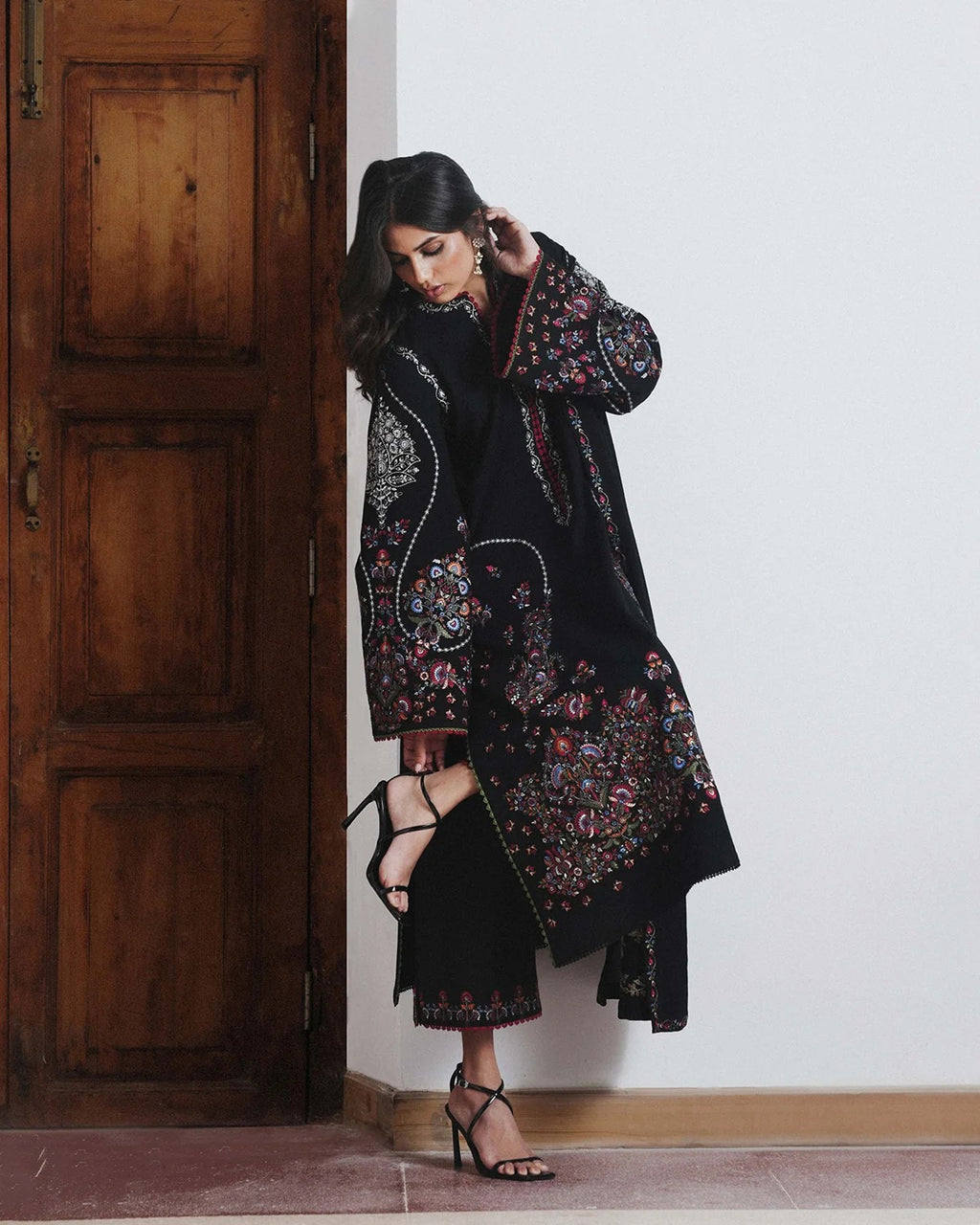 A woman models a long black embroidered kurta with colorful floral and paisley patterns, paired with black strappy high-heeled sandals, standing next to a rustic wooden door.