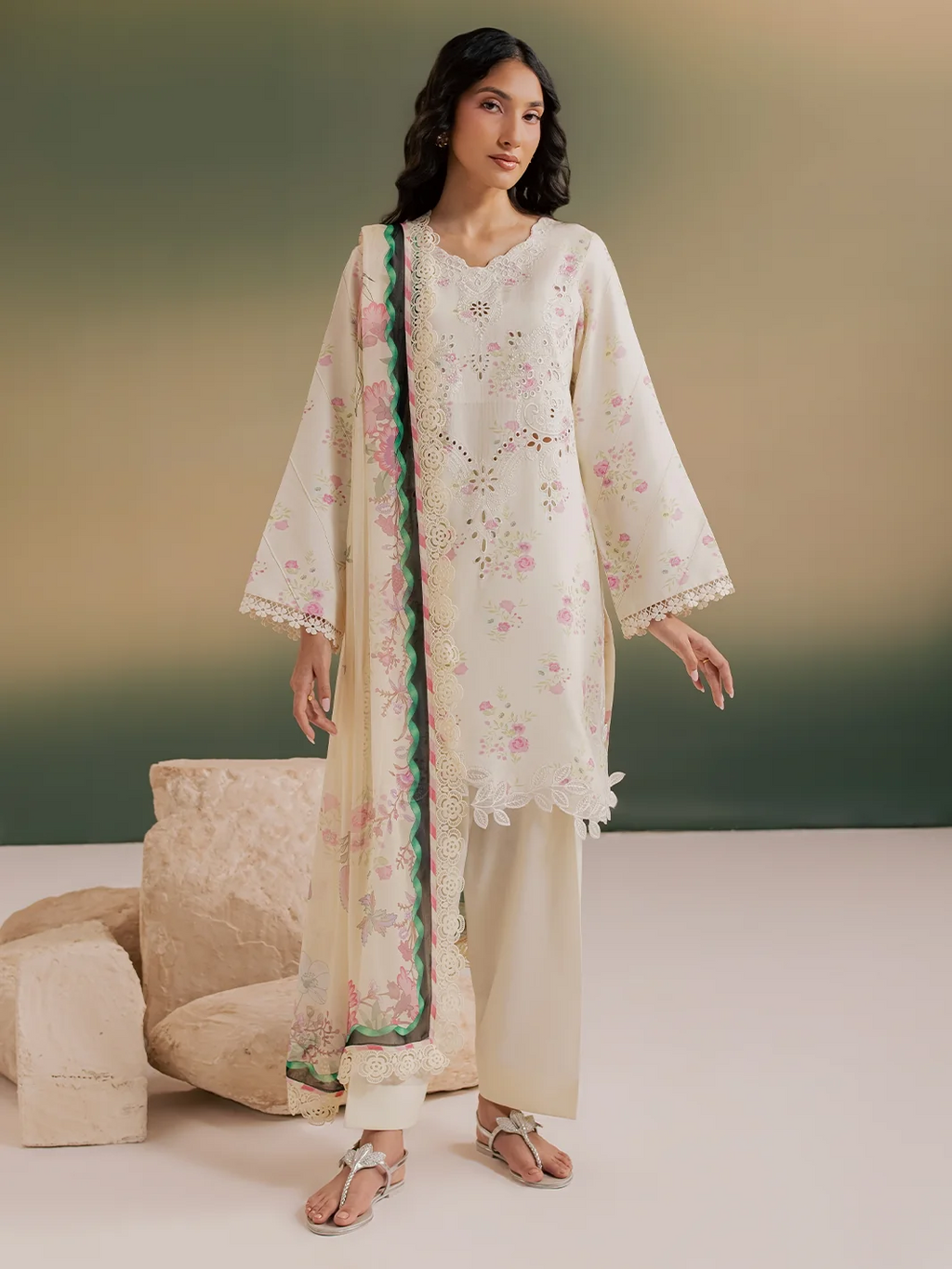 A woman models a traditional three-piece salwar kameez suit in cream with pink floral embroidery, featuring a matching dupatta with a green border, set against a neutral studio backdrop.