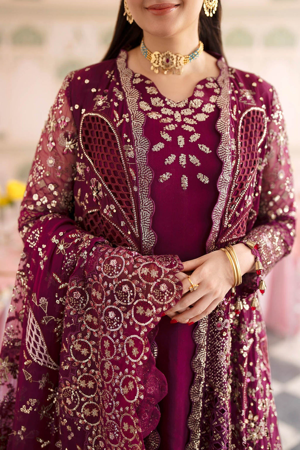 A close-up of a woman wearing an ornate deep magenta traditional outfit with intricate gold embroidery, a matching embroidered jacket, and a dupatta, accessorized with gold jewelry.