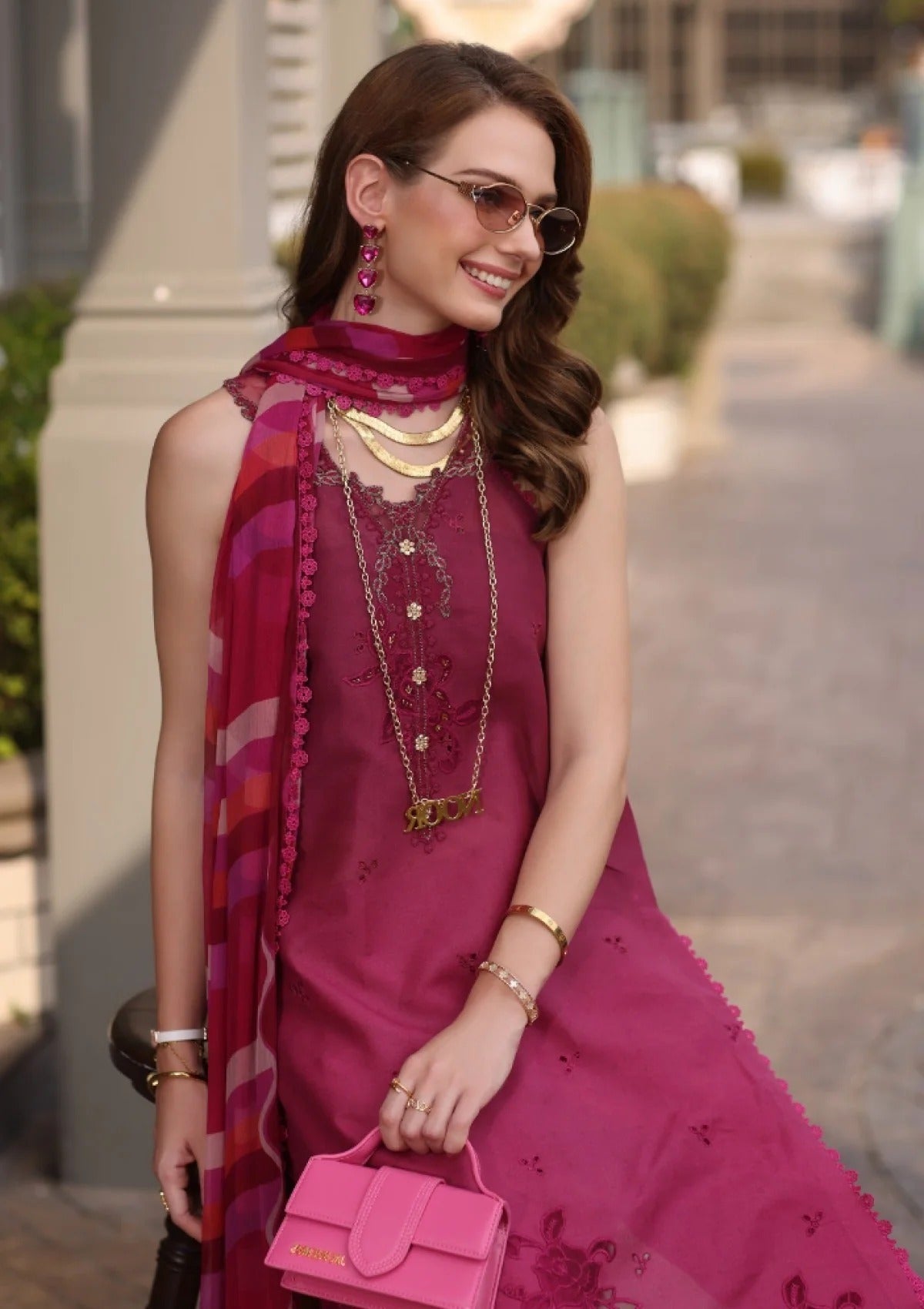 A woman wearing a magenta pink embroidered kurta with a matching scarf, layered gold necklaces, sunglasses, and holding a pink handbag.