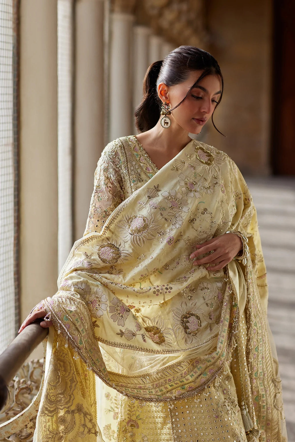 A woman wearing a light yellow embroidered saree with floral patterns and metallic detailing, standing in an elegant indoor corridor with columns.