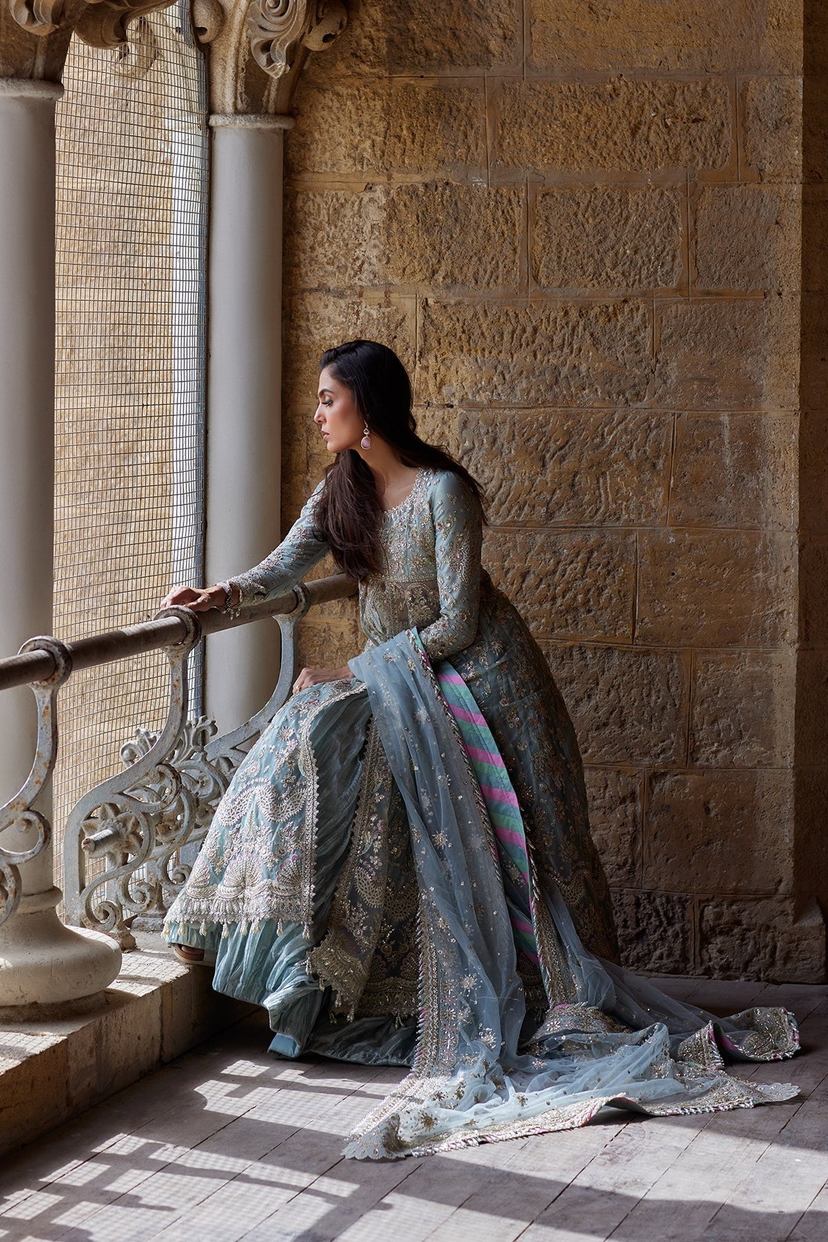 A woman wearing a traditional embroidered gown and dupatta sits on a stone balcony, looking out a window. The dress is a muted blue-gray with intricate silver and gold embroidery, and a bright pink and blue striped border detail is visible on the dupatta.