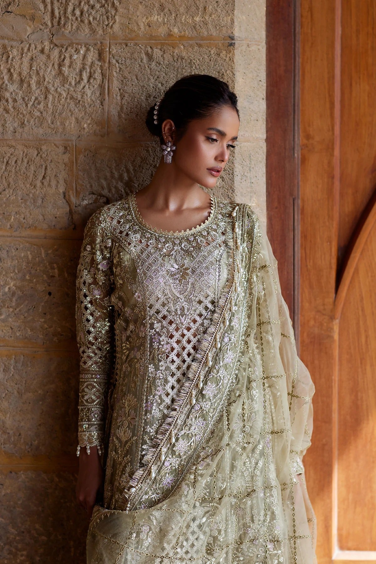 A woman wearing an ornate beige and gold embroidered traditional outfit with a sheer dupatta, standing against a stone wall and wooden door.
