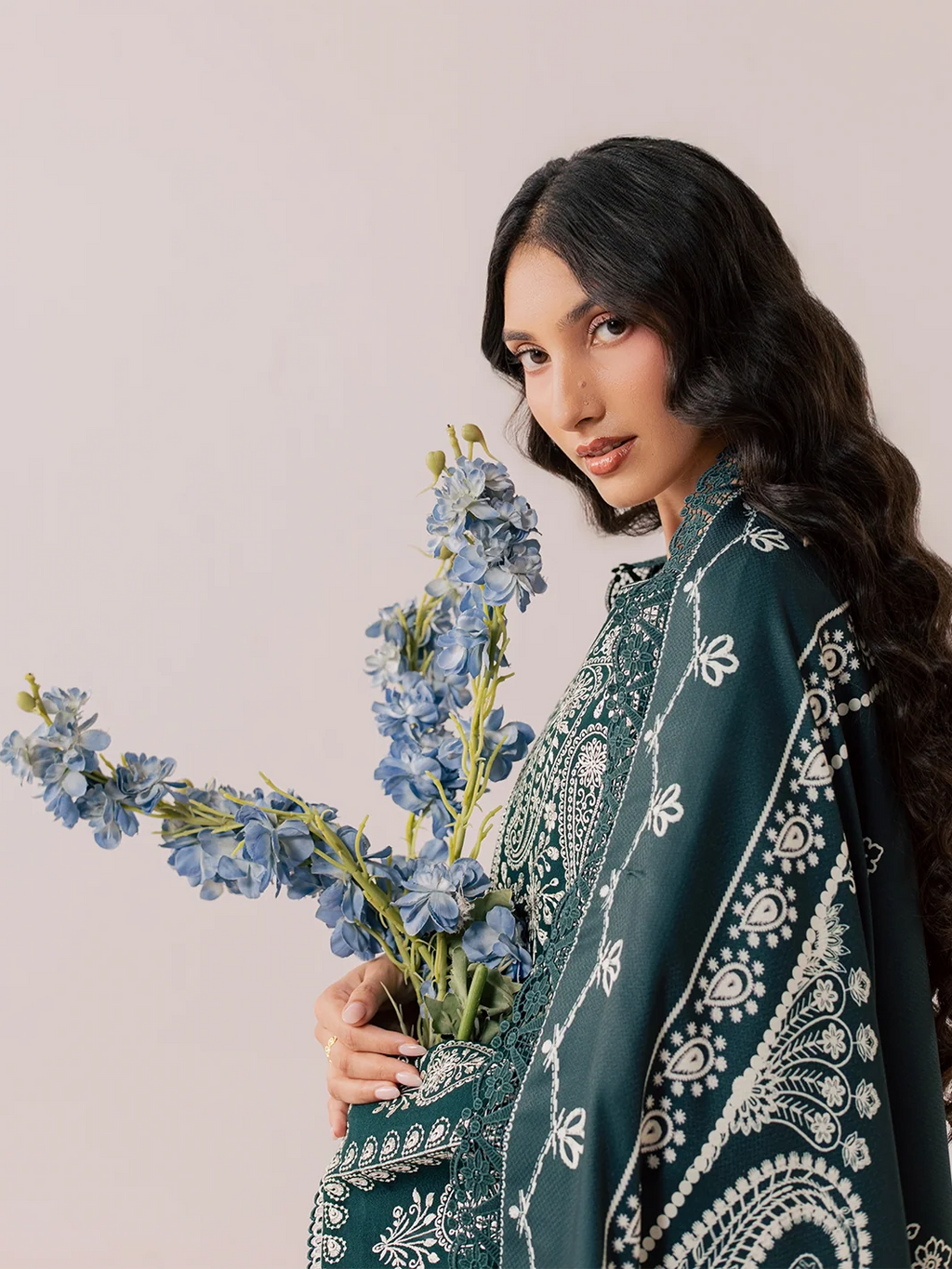 A woman modeling a dark teal green traditional outfit with intricate silver and white embroidery, featuring a matching embroidered dupatta draped over her shoulder against a light beige background.