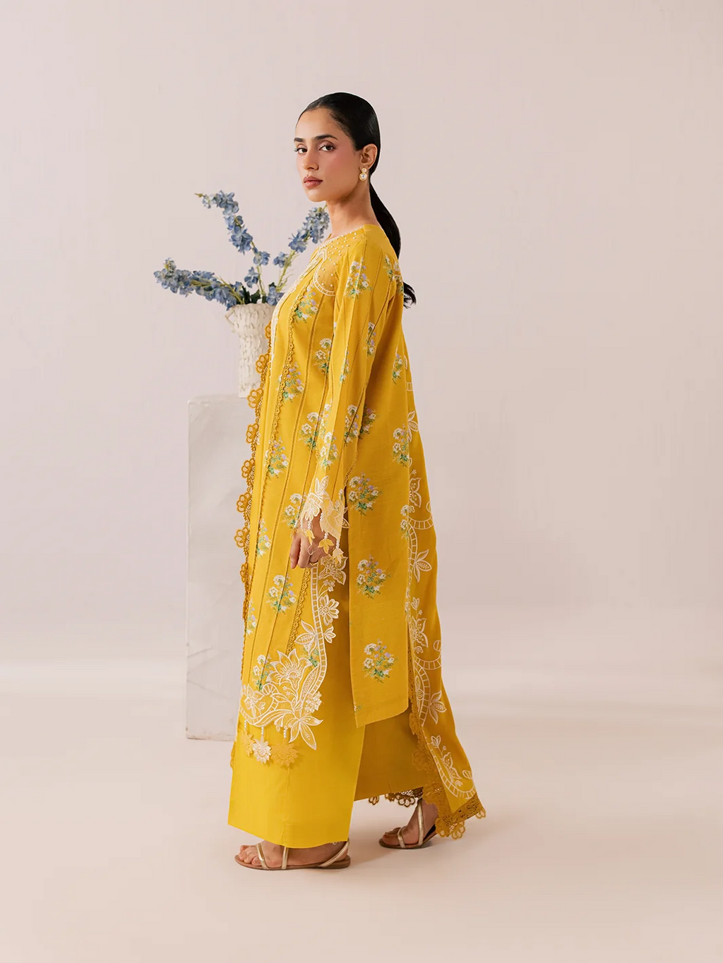 A woman modeling a bright yellow embroidered traditional outfit with a matching dupatta, standing in profile against a plain beige studio background.