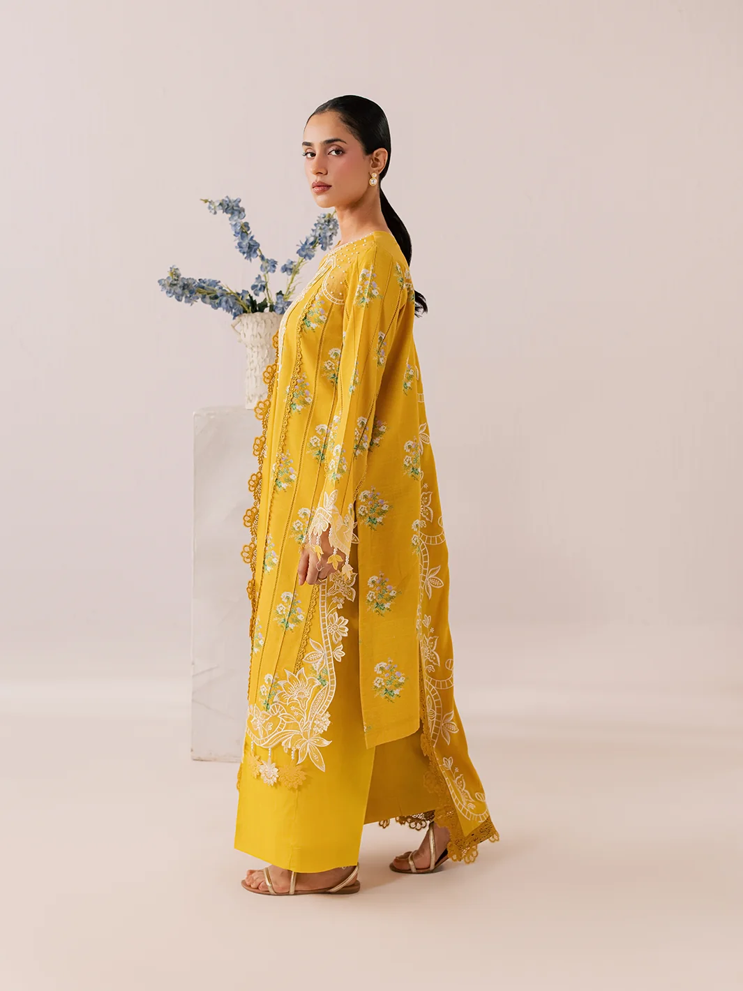 A woman modeling a bright yellow embroidered traditional outfit with a matching dupatta, standing in profile against a plain beige studio background.