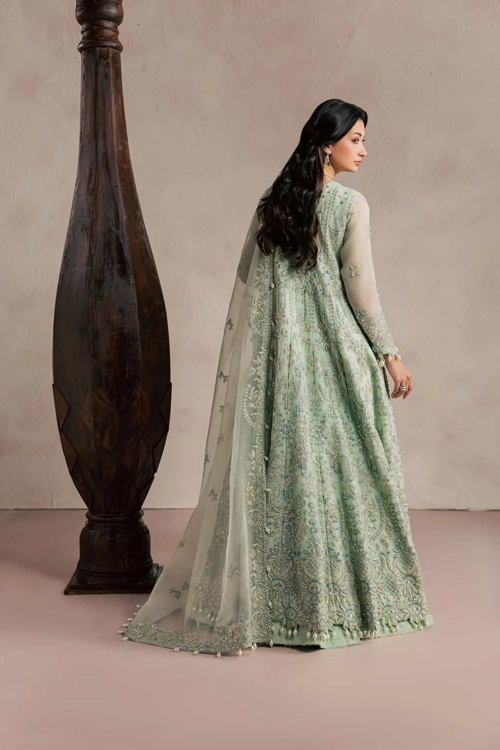 A woman wearing a light sage green embroidered Anarkali gown with sheer sleeves and a matching dupatta, standing beside a large dark wooden vase against a neutral beige studio backdrop.