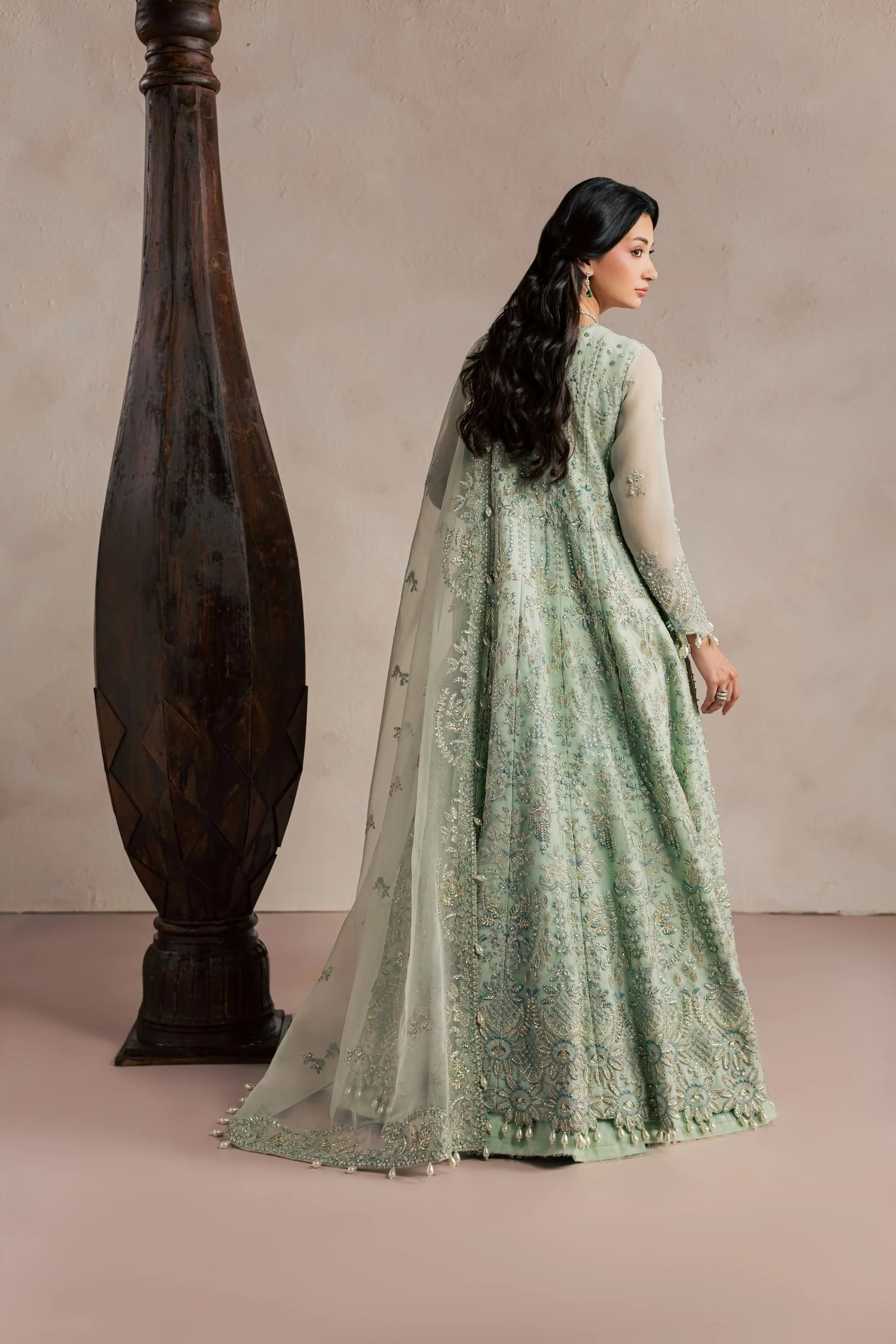 A woman wearing a light sage green embroidered Anarkali gown with sheer sleeves and a matching dupatta, standing beside a large dark wooden vase against a neutral beige studio backdrop.