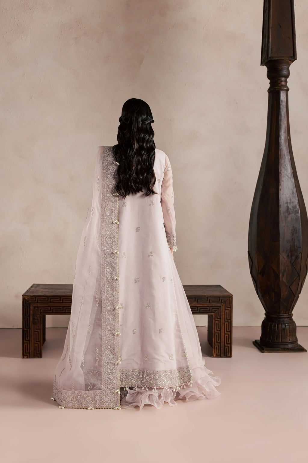 A woman viewed from behind wearing a traditional light pink or off-white long gown with embroidered details and a sheer draped overlay, standing in a styled interior with a bench and a dark wooden pillar against a warm beige wall.