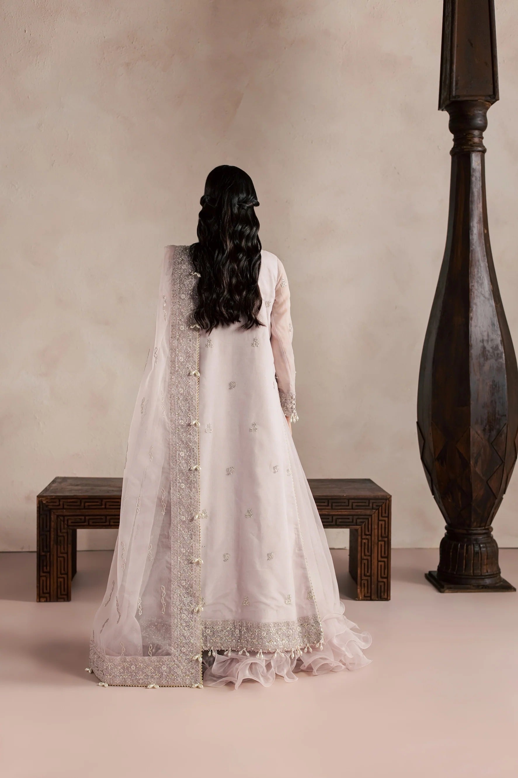 A woman viewed from behind wearing a traditional light pink or off-white long gown with embroidered details and a sheer draped overlay, standing in a styled interior with a bench and a dark wooden pillar against a warm beige wall.