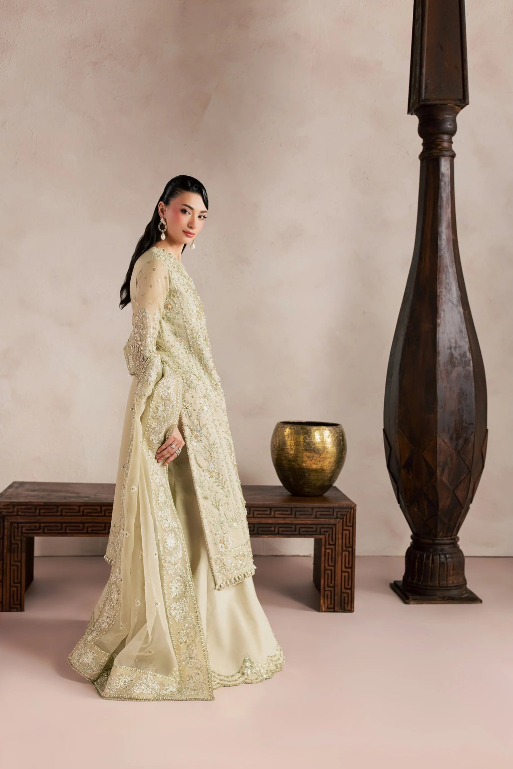 A woman models a traditional South Asian outfit in a warm beige studio setting with a wooden bench, a brass pot, and a tall dark wooden pillar. The outfit consists of a cream-colored embroidered kurta with a matching dupatta.