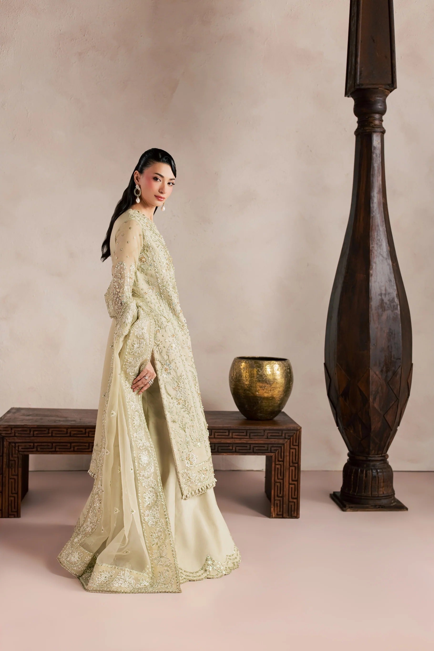 A woman models a traditional South Asian outfit in a warm beige studio setting with a wooden bench, a brass pot, and a tall dark wooden pillar. The outfit consists of a cream-colored embroidered kurta with a matching dupatta.