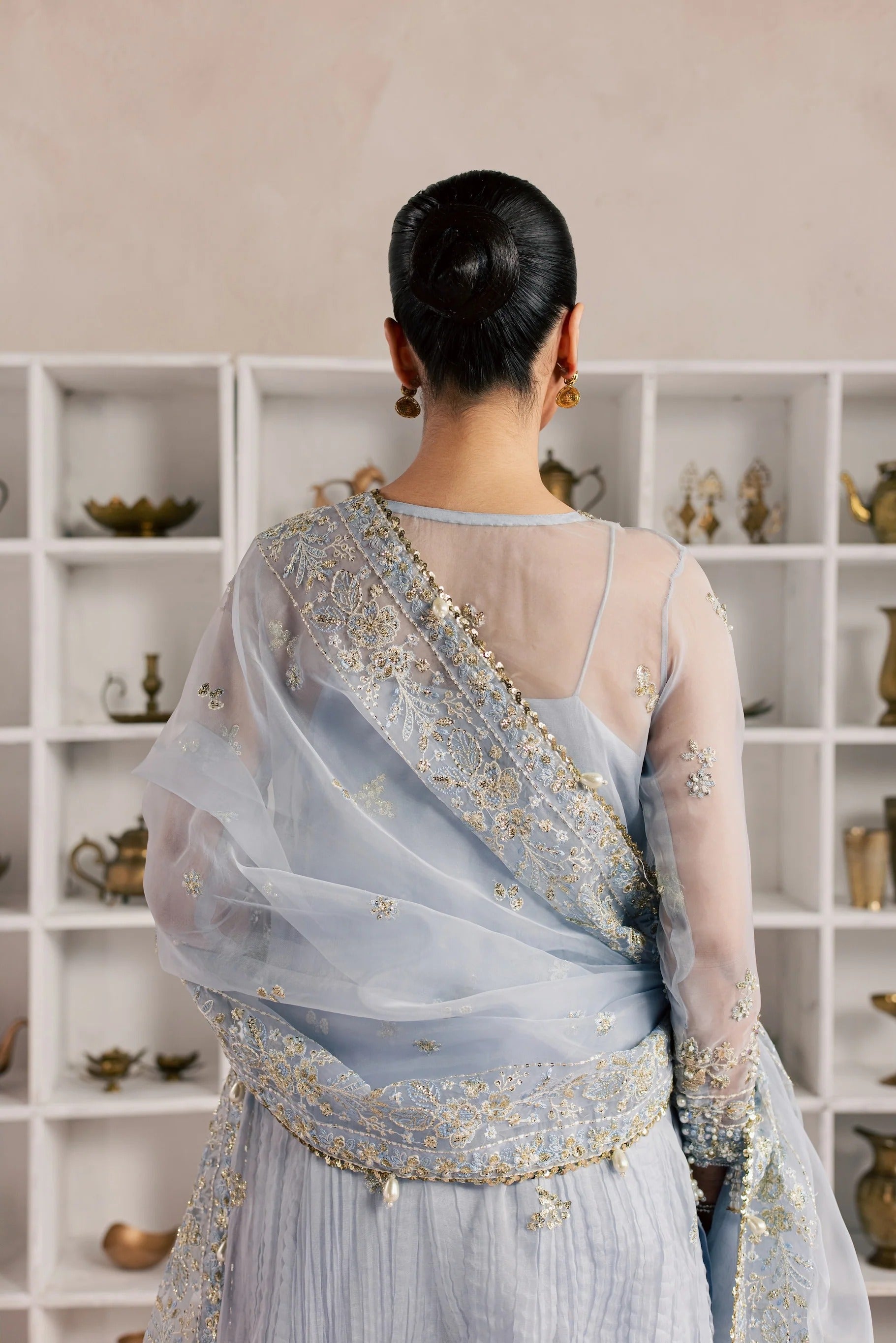 A woman viewed from behind wearing a light blue traditional outfit with gold embroidery and a sheer draped shawl, standing in front of white shelving.