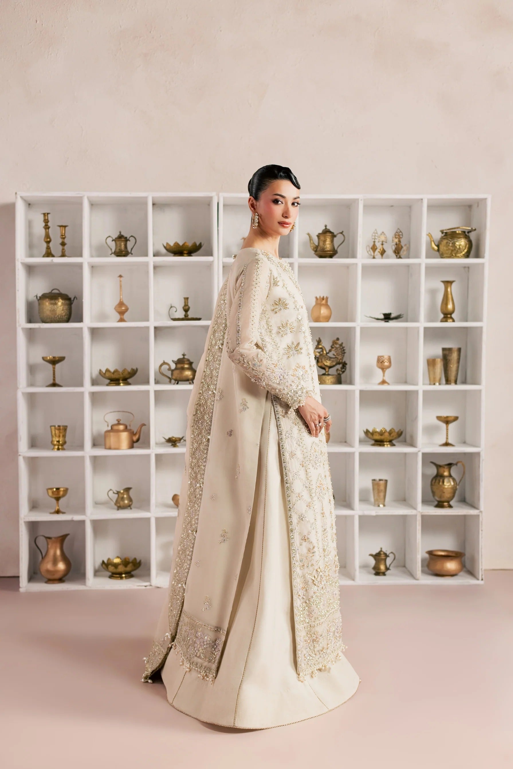 A woman models a traditional cream-colored embroidered gown with a matching sheer embroidered cape, standing in front of a white shelving unit filled with decorative brass and gold-toned vessels.