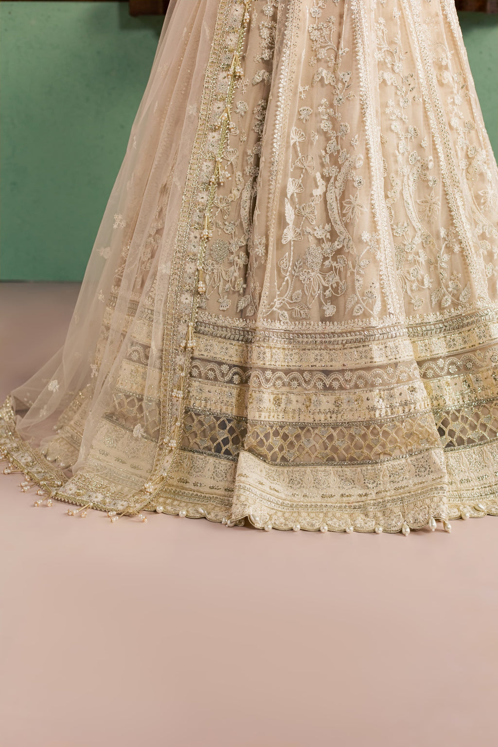 Close-up of a beige and gold heavily embroidered traditional skirt or lehenga with intricate lace, beadwork, and border detailing, shown against a soft pink floor and muted green wall.